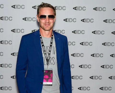 A man in sunglasses and a blue jacket stands in front of an AIDC backdrop.
