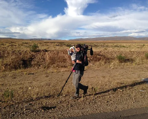 A person carries a film camera on a tripod across a dry plain.