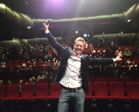 A man raises a microphone and opens his arms on a stage. A full cinema sits behind him.