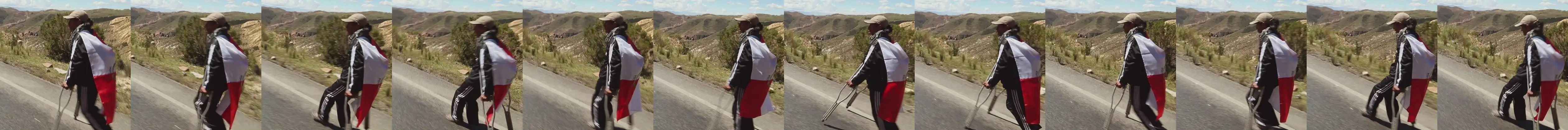 Frames of La Lucha: a man with a flag across his back walks along the highway with a crutch.