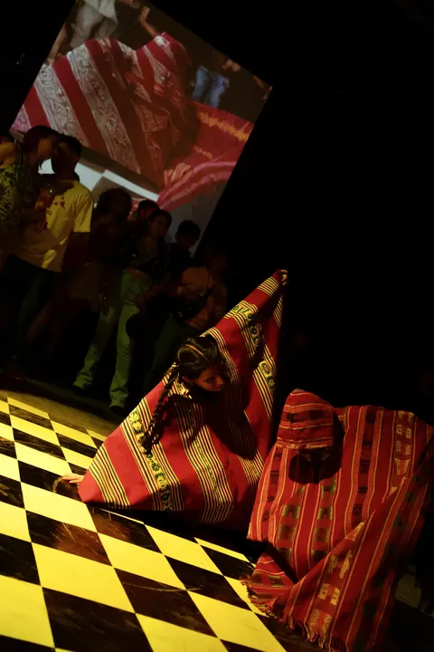 Two figures wrapped in red woven cloth crouch on a yellow and black checked floor.