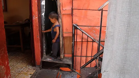 A small boy looks out of a doorway beside a metal staircase in an orange room. Still from Cocaine Prison.