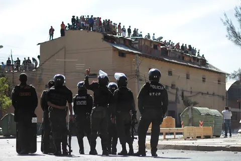 Riot police line up in front of a building whose roof is crowded with people. Still from Cocaine Prison.