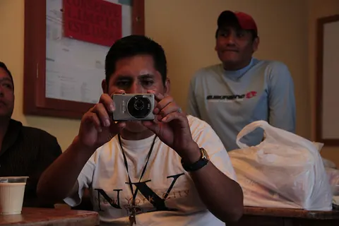A man holds up a small camera to film. A man in a red cap sits beside him. Still from Cocaine Prison.