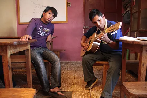 A man plays the guitar while another man sits and listens, in a room with a pink wall. Still from Cocaine Prison.