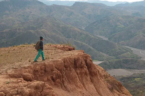 A person in green trousers stands on a red rocky ridge above deep valleys. Still from Cocaine Prison.
