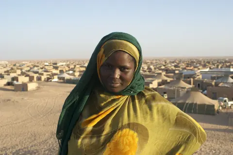 A woman in a green and yellow veil stands in front of a refugee camp in the desert. Still from Stolen.