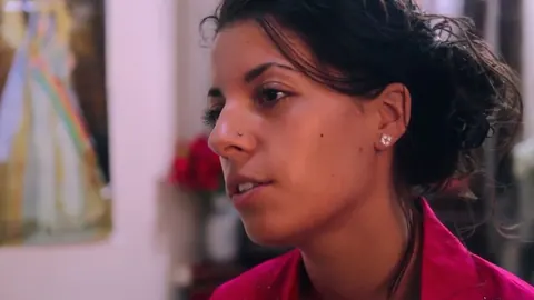 A young woman looks to one side in a room with a painting behind her. Still from The Bolivian Case.