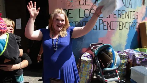 A woman in a blue dress waves outside a prison wall painted with lettering. Still from The Bolivian Case.