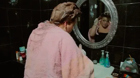 A woman in a pink towel faces a round mirror in a dark bathroom. Still from The Bolivian Case.