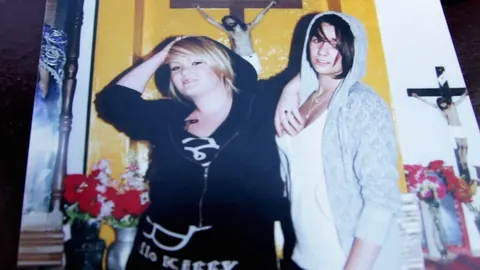 A photograph of two young women in hoodies, pinned up beside a crucifix and flowers. Still from The Bolivian Case.