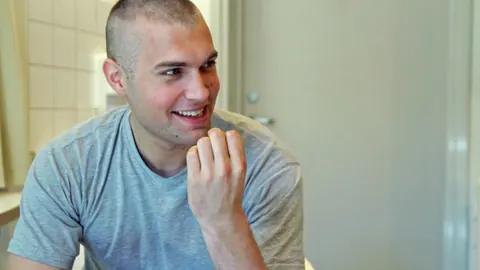 A young man with a shaved head smiles across a table. Still from The Bolivian Case.