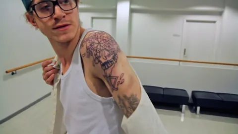 A young man in glasses and a vest shows a tattooed arm. Still from The Bolivian Case.