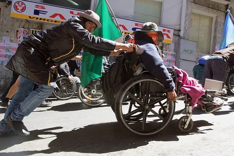 A man pushes a wheelchair up a steep street, a green flag behind him. Still from The Fight.