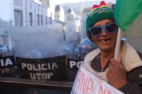 A smiling man in blue sunglasses and a woolly hat stands in front of police shields. Still from The Fight.