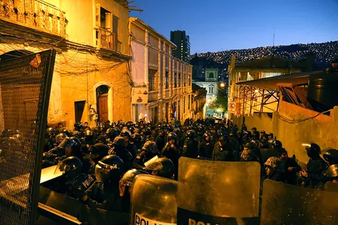 A crowd fills a narrow street at dusk, facing a line of police shields. Still from The Fight.