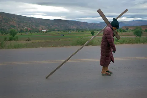 A person carries a wooden cross along an empty road between green fields. Still from The Fight.