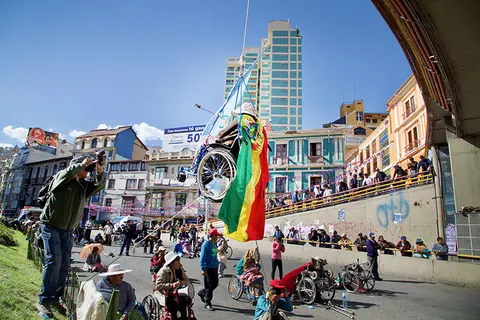 Protesters in wheelchairs gather in a street below a tall glass building, a Bolivian flag raised. Still from The Fight.
