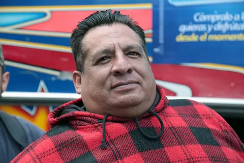 A man in a red checked hoodie looks at the camera in front of a bus. Still from The Fight.
