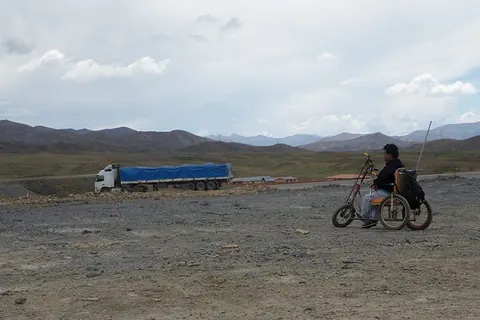 A person in a hand-powered wheelchair crosses a wide plain as a truck passes. Still from The Fight.