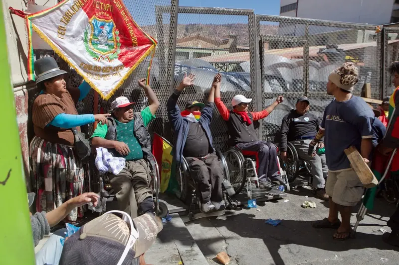 Protesters in wheelchairs raise their arms by a wire fence, under an embroidered banner.