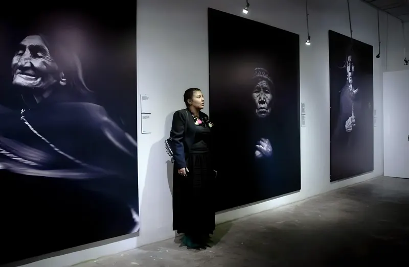 A woman in black stands in a gallery between large black-and-white portraits of Andean grandmothers.
