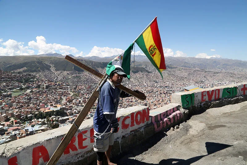 A man carries a wooden cross and a Bolivian flag on a hill above a city.