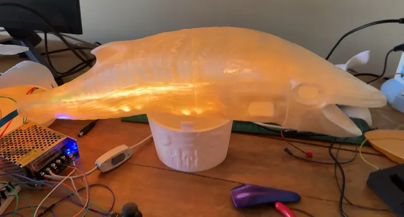 A translucent 3D-printed whale glows orange on a workbench, beside wires and a power supply.